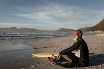 Homme âgé afro-américain sur la plage assis avec planche de surf regardant vers la mer. santé et bien-être, retraite active. — Photo de stock