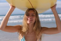 Улыбающаяся белая женщина в бикини с жёлтой доской для серфинга на голове на пляже. здоровый отдых на открытом воздухе у моря. — стоковое фото