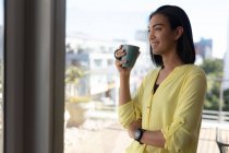 Smiling mixed race transgender woman standing on sunny roof terrace holding coffee. staying at home in isolation during quarantine lockdown. — Stock Photo