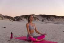 Mujer caucásica en la playa practicando yoga sentada en meditación. salud y bienestar, relajarse en la playa al amanecer. - foto de stock