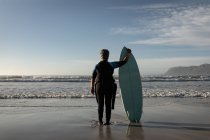 Visão traseira da mulher americana africana sênior segurando prancha de surf em pé na praia. viagem férias aposentadoria estilo de vida conceito — Fotografia de Stock