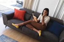 Смешанная расовая трансгендерная женщина отдыхает в гостиной, сидя на диване и читая книгу. оставаться дома в изоляции во время карантинной изоляции. — стоковое фото