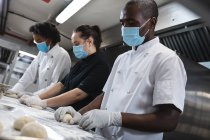 Mixed race professional chefs preparing dough wearing sanitary gloves and face mask. working in a busy restaurant kitchen during coronavirus covid 19 pandemic. — Stock Photo