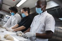 Gemischte Profiköche bereiten Teig mit Hygienehandschuhen und Mundschutz zu. Arbeit in einer belebten Restaurantküche während der Coronavirus-Pandemie 19. — Stockfoto