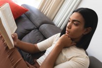 Transgender-Frau mit gemischter Rasse entspannt sich im Wohnzimmer und sitzt auf der Couch und liest Buch. Isolationshaft während der Quarantäne. — Stockfoto