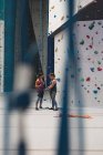 Instrutor feminino caucasiano que mostra à mulher como nó uma corda a um arnês na parede de escalada interior. fitness e tempo de lazer no ginásio. — Fotografia de Stock