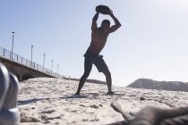 African american man exercising with weights on beach on sunny day. healthy outdoor lifestyle fitness training. - foto de stock