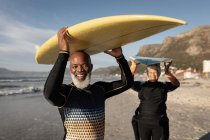 Retrato de una pareja afroamericana mayor llevando tabla de surf en la cabeza en la playa. viaje vacaciones retiro estilo de vida concepto - foto de stock