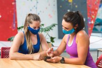 Dos mujeres caucásicas felices en máscaras faciales usando un teléfono inteligente en la pared de escalada interior. fitness y tiempo libre en el gimnasio durante coronavirus covid 19 pandemia. - foto de stock