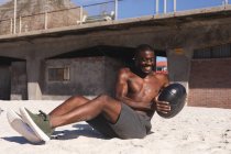 Усміхнений афроамериканський чоловік тренується з вагами на пляжі в сонячний день. здорова фітнес-тренування на відкритому повітрі . — стокове фото