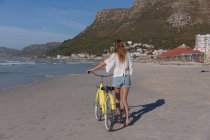 Rear view of caucasian woman in white top and shorts walking with a bicycle at the beach. healthy outdoor leisure time by the sea. — Photo de stock