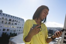 Mujer transexual de raza mixta sonriente de pie en la terraza soleada de la azotea sosteniendo el café con un teléfono inteligente. permanecer en casa aislado durante el bloqueo de cuarentena. - foto de stock