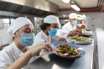 Diversa raza de chefs profesionales masculinos y femeninos regalando platos con máscaras faciales. trabajando en una ajetreada cocina de restaurante durante coronavirus covid 19 pandemia. - foto de stock