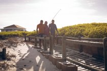 African american father and his two sons with fishing rods walking together on the bridge. summer beach holiday and leisure concept. — Stock Photo