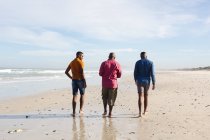 Vista trasera del padre afroamericano y sus dos hijos caminando juntos en la playa. vacaciones de playa de verano y concepto de ocio. - foto de stock