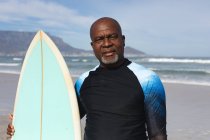 Retrato de un hombre mayor afroamericano con tabla de surf en la playa. vacaciones de playa de verano y concepto de ocio. - foto de stock