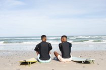 Vista trasera de hermanos afroamericanos con tablas de surf sentados en la playa. vacaciones de playa de verano y concepto de ocio. - foto de stock