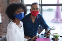 Homme métis souriant et femme afro-américaine portant un masque assis, en utilisant une tablette dans un café. créatifs numériques sur la route pendant coronavirus covid 19 pandémie. — Photo de stock