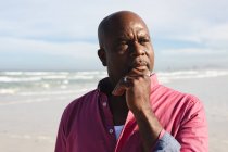 Afrikanischer älterer Mann mit der Hand am Kinn, der am Strand steht. Sommer-Strandurlaub und Freizeitkonzept. — Stockfoto
