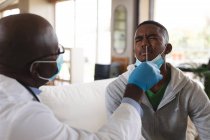 Medico afroamericano senior maschio che esegue test nasali su un uomo afroamericano a casa. test medici per la prevenzione del concetto di focolaio di coronavirus — Foto stock