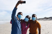 Der afroamerikanische Vater und seine beiden Söhne tragen Gesichtsmasken und machen gemeinsam ein Selfie am Strand. Sommerurlaub am Strand während des Covid-19-Pandemiekonzepts. — Stockfoto