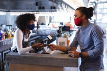 Diverse männliche und weibliche Kellner tragen Gesichtsmasken und benutzen Zahlungsterminals im Café. Unabhängiges Café, Geschäfte während der Coronavirus-Pandemie 19. — Stockfoto