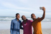 Le père afro-américain et ses deux fils prennent un selfie sur un smartphone à la plage. vacances à la plage d'été et concept loisirs. — Photo de stock