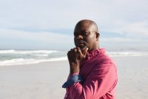 Hombre mayor afroamericano con la mano en la barbilla de pie en la playa. vacaciones de playa de verano y concepto de ocio. - foto de stock