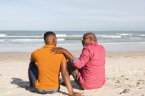 Padre afroamericano che parla con suo figlio seduto in spiaggia. estate vacanza al mare e concetto di svago. — Foto stock