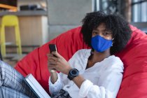 Afroamerikanerin mit Mundschutz, sitzend, Smartphone im Café. Digitale Kreative unterwegs während der Coronavirus-Pandemie. — Stockfoto