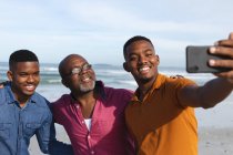 El padre afroamericano y sus dos hijos tomando un selfie de un smartphone en la playa. vacaciones de playa de verano y concepto de ocio. - foto de stock