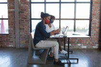 Femme afro-américaine et homme métis portant des masques assis dans un café à l'aide d'un ordinateur portable. créatifs numériques sur la route pendant coronavirus covid 19 pandémie. — Photo de stock
