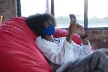 Mujer afroamericana usando mascarilla, sentada, usando smartphone en la cafetería. creativos digitales en movimiento durante la pandemia del coronavirus covid 19. - foto de stock