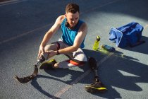 Atleta maschio caucasico che fissa la gamba protesica mentre è seduto sulla pista di corsa nello stadio. concetto di sport paralimpico — Foto stock