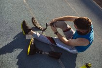 Atleta maschio caucasico che fissa la gamba protesica mentre è seduto sulla pista di corsa nello stadio. concetto di sport paralimpico — Foto stock