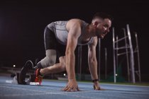 Atleta masculino caucasiano com perna protética em posição inicial para correr na pista à noite. conceito de esporte paralímpico — Fotografia de Stock