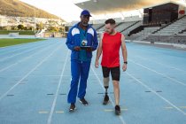 Treinador masculino afro-americano instruindo atleta masculino caucasiano com perna protética na pista de corrida. conceito de esporte paralímpico — Fotografia de Stock