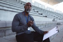 Entrenador masculino afroamericano con cronómetro midiendo el tiempo mientras está sentado en los asientos del estadio. concepto de deporte paralímpico - foto de stock