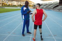Entrenador masculino afroamericano instruyendo a atleta masculino caucásico con pierna protésica en pista de atletismo. concepto de deporte paralímpico - foto de stock