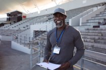 Retrato del entrenador afroamericano que sostiene el portapapeles sonriendo mientras está de pie en el estadio. concepto de deporte paralímpico - foto de stock