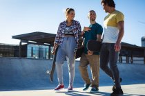 Tres amigas caucásicas felices caminando con monopatines y hablando al sol. pasando el rato en un skatepark urbano en verano. - foto de stock