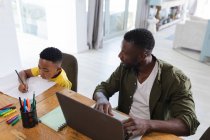 Pai e filho afro-americanos sentados à mesa, usando laptop e escrevendo em notebook. em casa em isolamento durante o confinamento de quarentena. — Fotografia de Stock