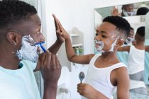 Pai e filho afro-americanos na casa de banho, a barbear-se juntos. em casa em isolamento durante o confinamento de quarentena. — Fotografia de Stock