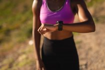 Sección media de la mujer afroamericana en forma comprobando smartwatch durante el ejercicio en el campo. estilo de vida activo saludable y fitness al aire libre. - foto de stock