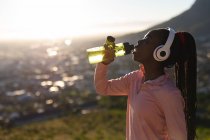 African american woman drinking water, taking break in exercise outdoors, wearing headphones. healthy active lifestyle and outdoor fitness. — Stock Photo