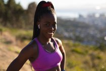 Портрет усміхненої афроамериканської жінки з бездротовими навушниками, займається в сільській місцевості. здоровий активний спосіб життя та фізична підготовка на відкритому повітрі . — стокове фото