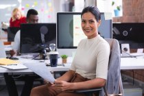 Retrato de mulher indiana segurando um documento sorrindo enquanto se senta em sua mesa no escritório moderno. negócio, profissionalismo e conceito de escritório — Fotografia de Stock