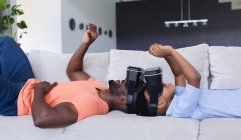 Африканский американец отец и сын лежат на диване в наушниках и трогают виртуальный экран. в доме в изоляции во время карантинной изоляции. — стоковое фото