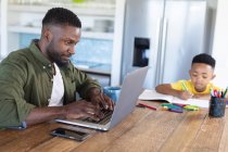Afroamerikanische Vater und Sohn sitzen am Tisch, benutzen Laptop und schreiben in Notizbuch. Zuhause in Isolation während der Quarantäne. — Stockfoto