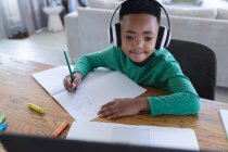 Niño afroamericano en la clase de la escuela en línea, usando auriculares y portátil. en casa en aislamiento durante el bloqueo de cuarentena. - foto de stock